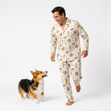 A man wearing a matching long-sleeve pajama set with a festive cartoon print walks barefoot while smiling, accompanied by a small corgi dog on a seamless white studio background.