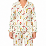 A model wearing a matching long-sleeve button-up pajama top and pants set in cream, covered in a festive holiday print featuring teddy bears in Santa hats, Christmas trees, ornaments, and the word 'Oogie Oogie'.
