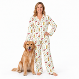 A woman wearing a matching long-sleeve pajama set with a colorful Santa-themed print stands next to a golden retriever dog on a plain white studio background.