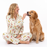 A woman in a festive Christmas-themed pajama set sits cross-legged on a white studio floor, smiling as she offers a treat to a golden retriever dog. The pajamas feature a cream background with a colorful print of Santa Claus, cookies, and holiday motifs.