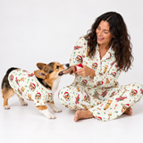 A woman and a corgi dog wearing matching Christmas-themed pajamas sit together on a white studio background. The pajamas have an off-white base with a festive print of Santa clowns, gingerbread cookies, ornaments, and holiday motifs.