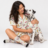 A woman and a Dalmatian dog sit together on a white studio floor wearing matching holiday-themed pajamas with a cartoon dog print, Santa hats, and red accents. The woman also wears matching knee-high socks.