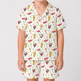 A child wearing a matching short-sleeve collared pajama top and shorts set with a festive Christmas-themed print featuring Santa clowns, Christmas trees, ornaments, and red sleighs on an off-white background, photographed against a light gray studio backdrop.