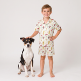 A young boy stands barefoot next to a black, white, and tan dog, wearing a matching short-sleeve button-up pajama shirt and shorts set with a colorful cartoon print on a cream background, photographed against a light gray studio backdrop.