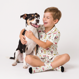A young boy wearing a matching short-sleeve pajama shirt and shorts set with a colorful cartoon dog print sits cross-legged on a white studio floor, hugging a black, white, and tan dog. The pajamas have a cream background with multicolor cartoon dog illustrations.