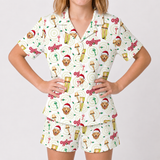 A woman modeling a matching short-sleeve pajama set with a white base and an all-over festive print featuring a golden retriever, beer glasses, Christmas hats, and red text on a light gray studio background.