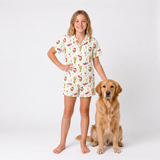 A young girl stands barefoot next to a golden retriever dog wearing a matching short-sleeve pajama set with a colorful cartoon food and drink print on a white background.