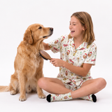 A young girl with light brown hair sits cross-legged on a white studio floor, smiling as she brushes a golden retriever dog. The girl is wearing a matching pajama set with a white background and a colorful holiday-themed print, including Santa Claus, reindeer, and snowflakes.
