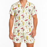 A man wearing a matching short-sleeve button-up pajama shirt and shorts set with a festive holiday-themed print featuring Christmas trees, drinks, Santa hats, and the word 'Odds'.