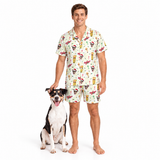 A man wearing a matching short-sleeve pajama shirt and shorts set with a colorful Santa Claus-themed print, standing next to a black and white dog on a solid white background.