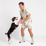 A man wearing a matching short-sleeve button-up pajama shirt and shorts set with a dog-themed print, standing in a playful pose with a black and white dog on a white studio background.
