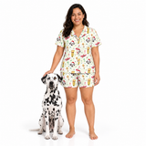 A woman stands barefoot next to a Dalmatian dog on a white studio background, wearing a matching short-sleeve pajama set with a white base and a colorful cartoon print featuring dogs, drinks, and festive motifs.