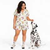 A woman wearing a matching pajama set with a Dalmatian dog sitting beside her, all featuring a festive holiday print with Santa Claus dogs, snow globes, and hot air balloons on a white background.
