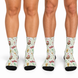 Three pairs of crew socks shown on lower legs against a white background. The socks have a light cream base with a festive pattern featuring Santa Claus faces, Christmas trees, ornaments, and the word 'Merry' in red, green, and gold, with solid black toes and heels.
