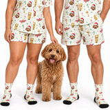 A couple wearing matching holiday-themed pajamas and socks stands next to a small fluffy dog on a white studio background. The pajama set has a cream base with a repeating print of a brown dog wearing a Santa hat, beer glasses, and Christmas-themed illustrations.