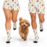 A person in matching white patterned shorts and socks walks alongside a small fluffy brown dog on a clean white studio background. The socks and shorts feature a holiday-themed print with Santa hats, gingerbread cookies, candy canes, and drinks.