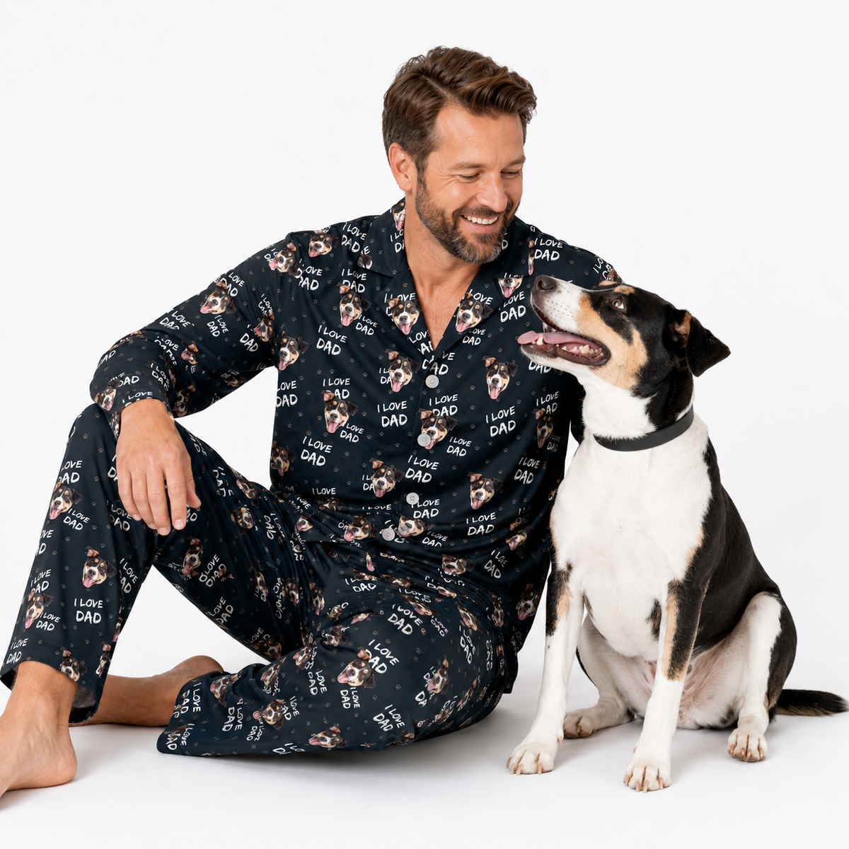 Man wearing a pajama set with dog prints sitting next to a dog on a white background