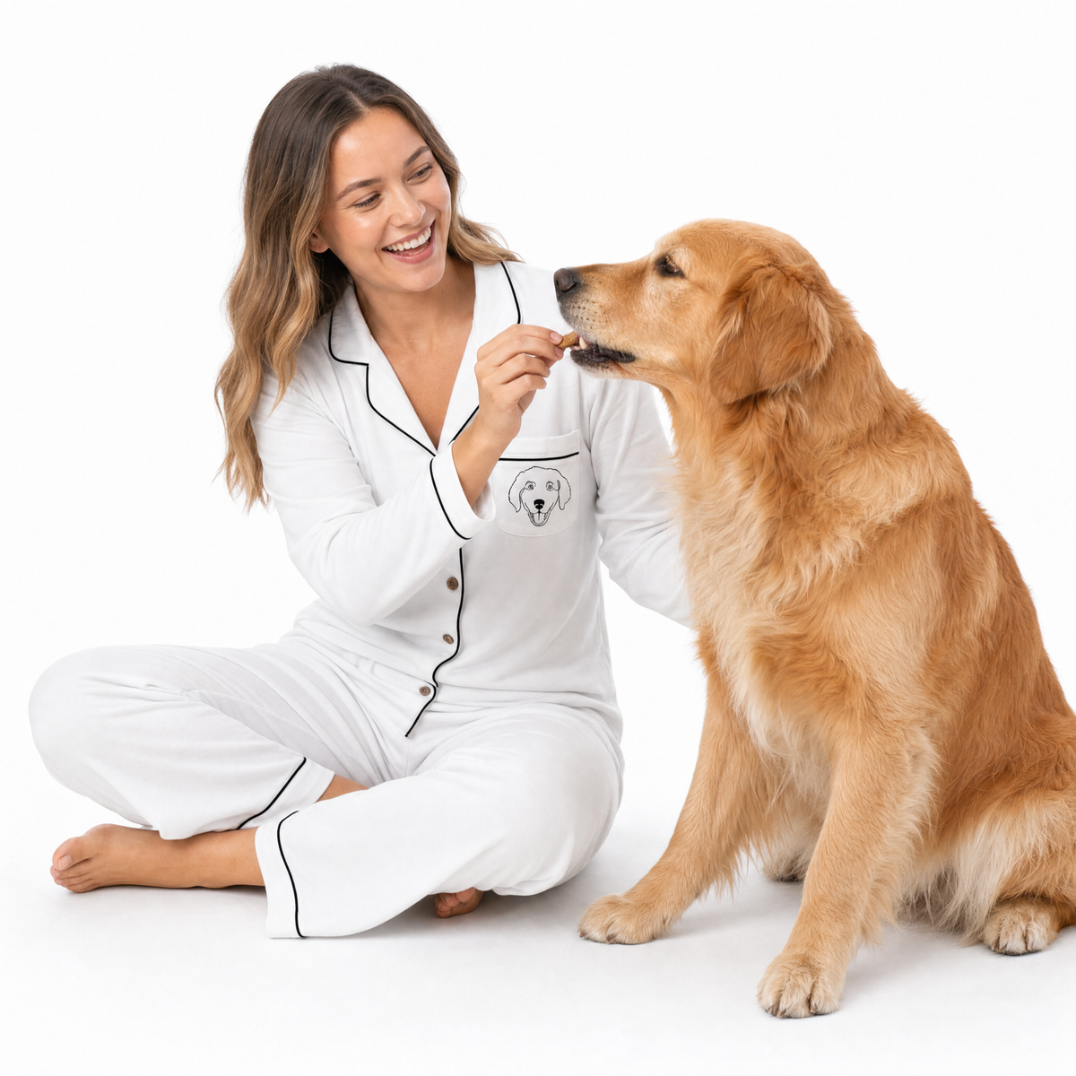Woman in ivory pajamas feeding a treat to a golden retriever on a white background