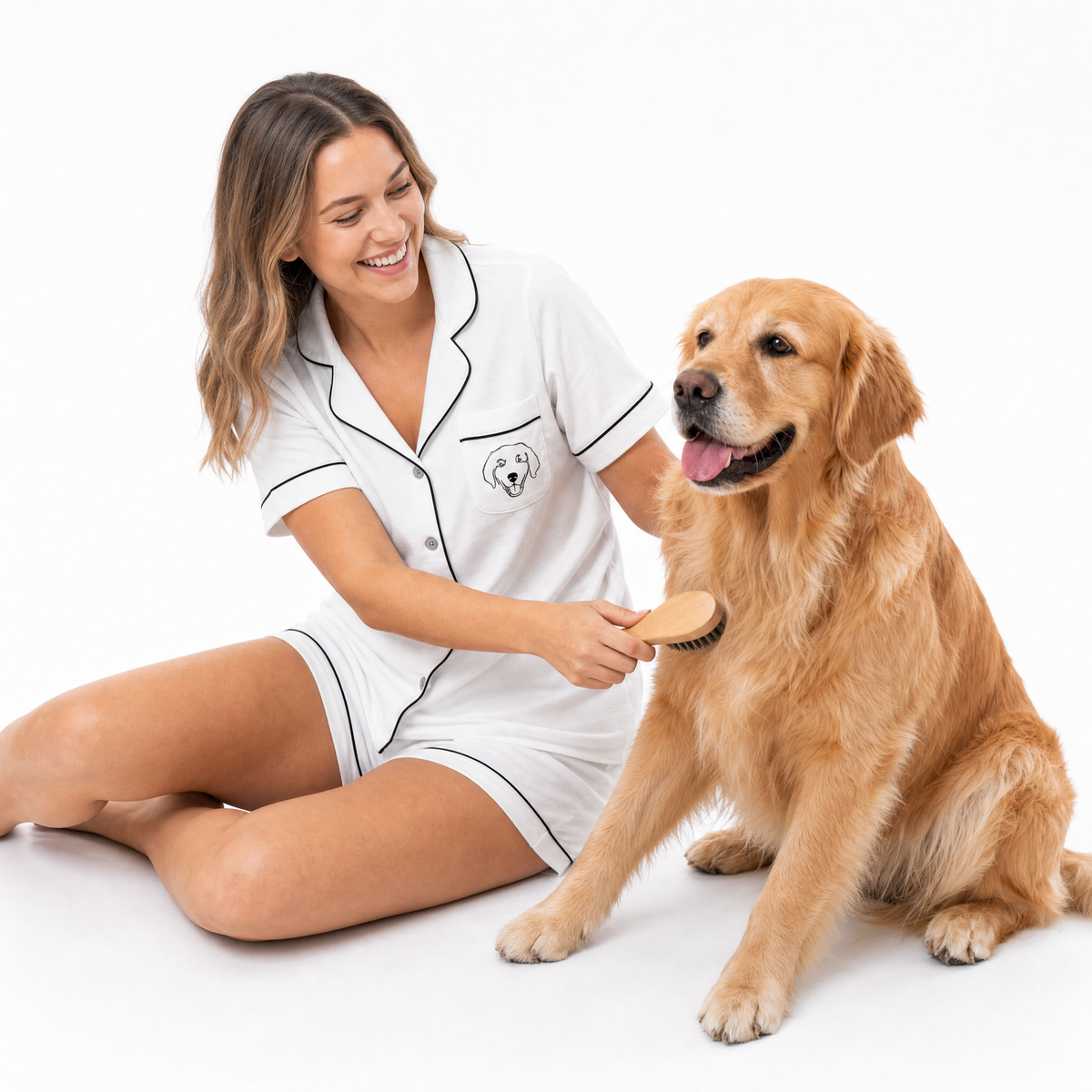 Woman in a white pajama set brushing a golden retriever on a white background