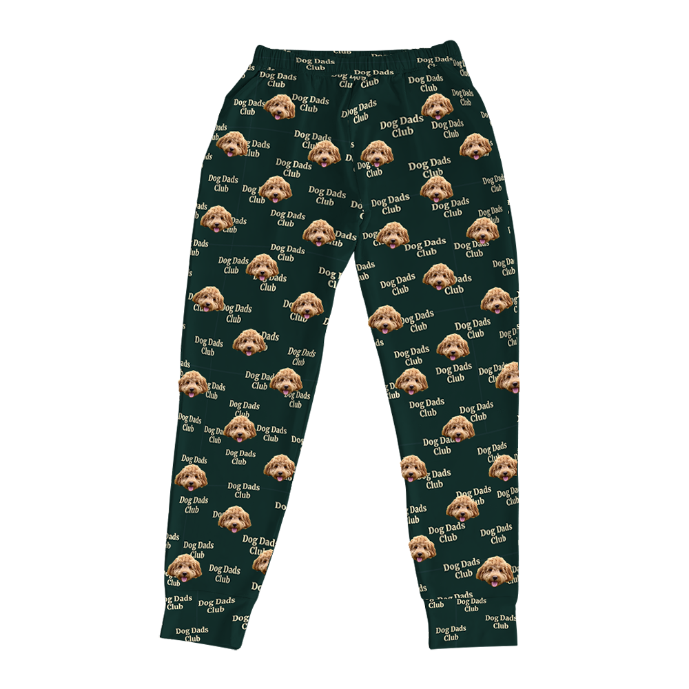 Hunter green jogger pajama pants with an all-over pattern featuring a brown curly-haired dog’s face and the repeated text ‘Dog Dads Club’ in beige, displayed flat on a white background.