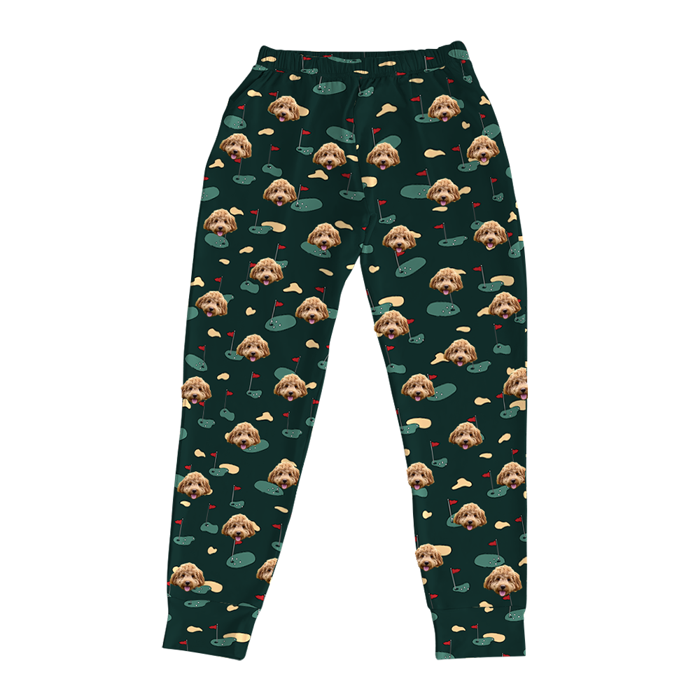 Dark green pajama joggers covered in a repeating golf-themed pattern. The pattern features alternating elements: cartoon depictions of golf putting greens with a red flag, yellowish sand traps, and smiling headshots of a light brown dog.