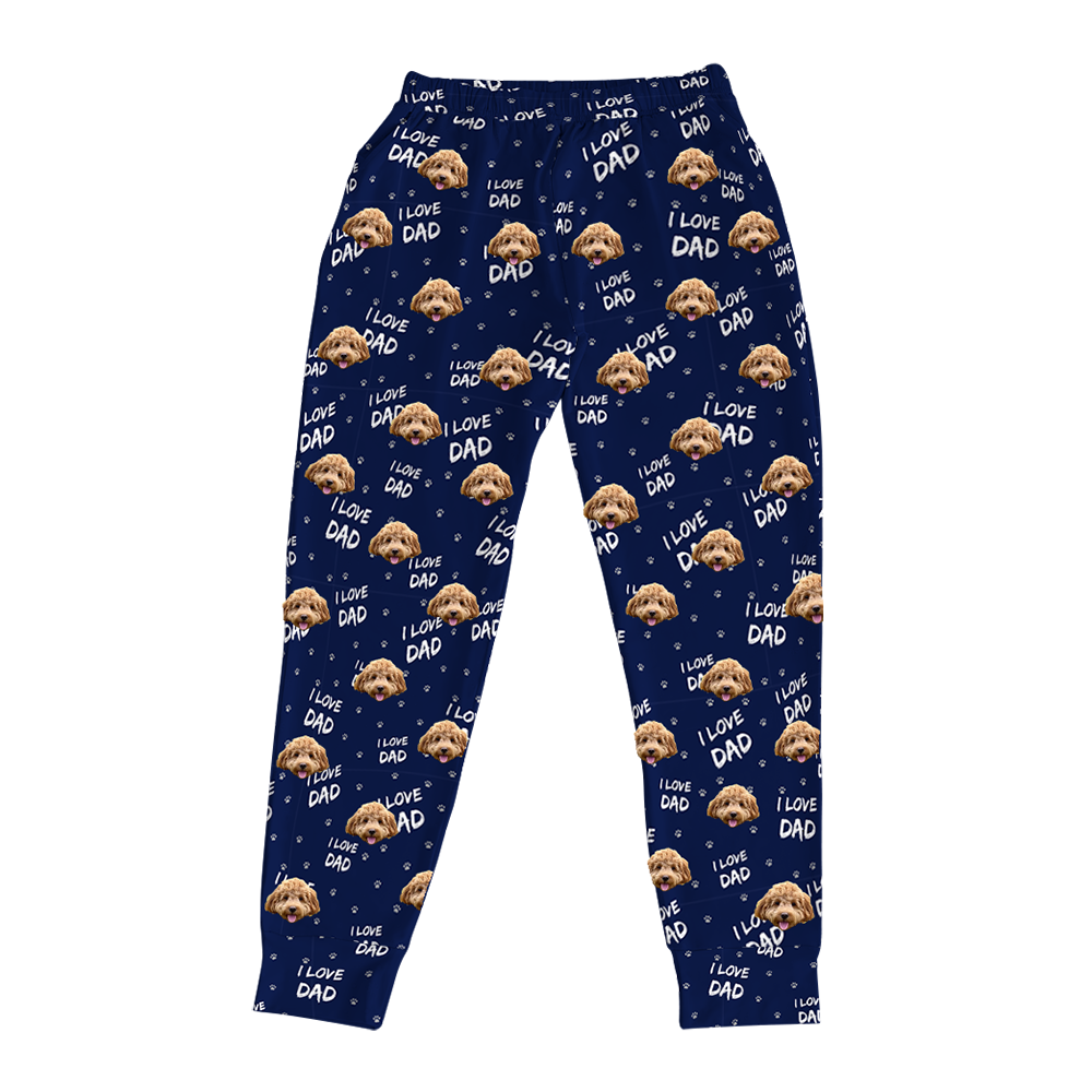 Navy blue jogger pajama pants featuring an all-over print of a brown curly-haired dog’s face, white paw prints, and the repeated text ‘I LOVE DAD,’ displayed flat on a white background.