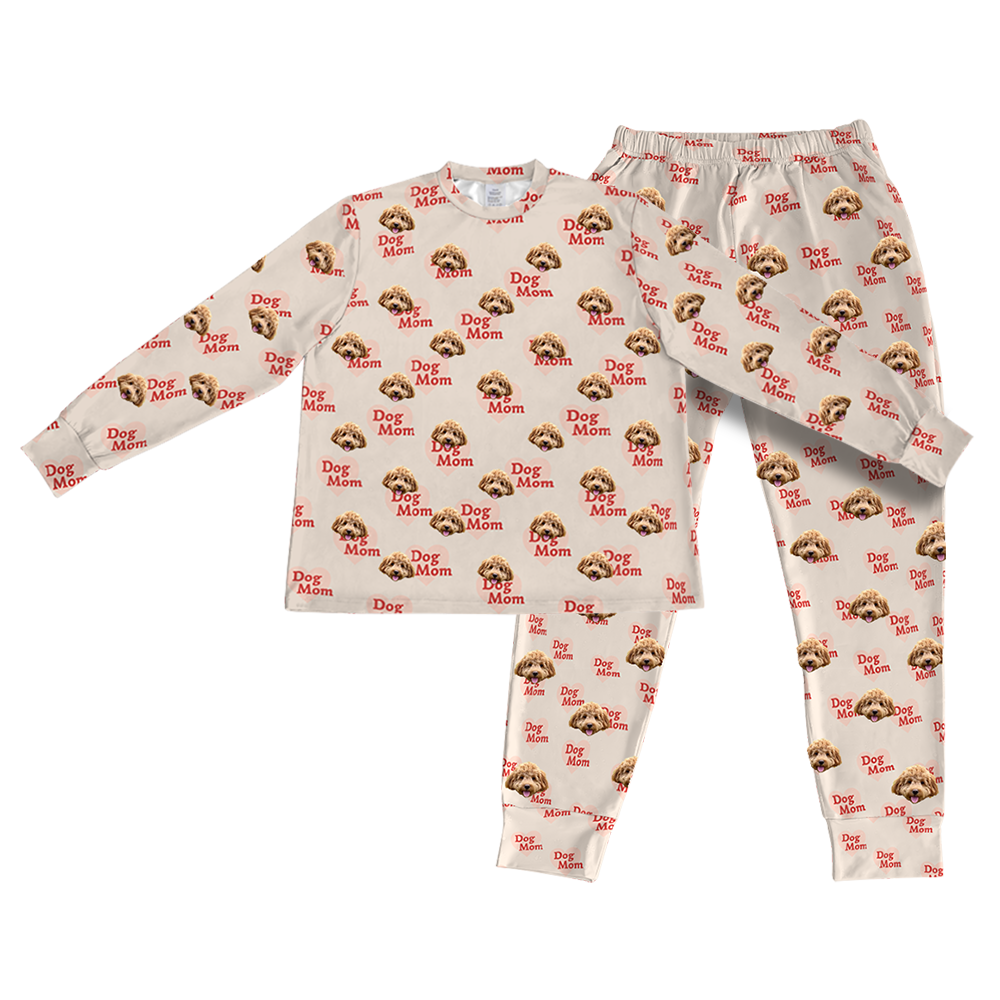 A matching set of long-sleeve pajama top and jogger bottoms in a light beige fabric. The entire set is covered in a repeating pattern featuring alternating elements: the phrase "Dog Mom" in red text and smiling headshots of a dog.