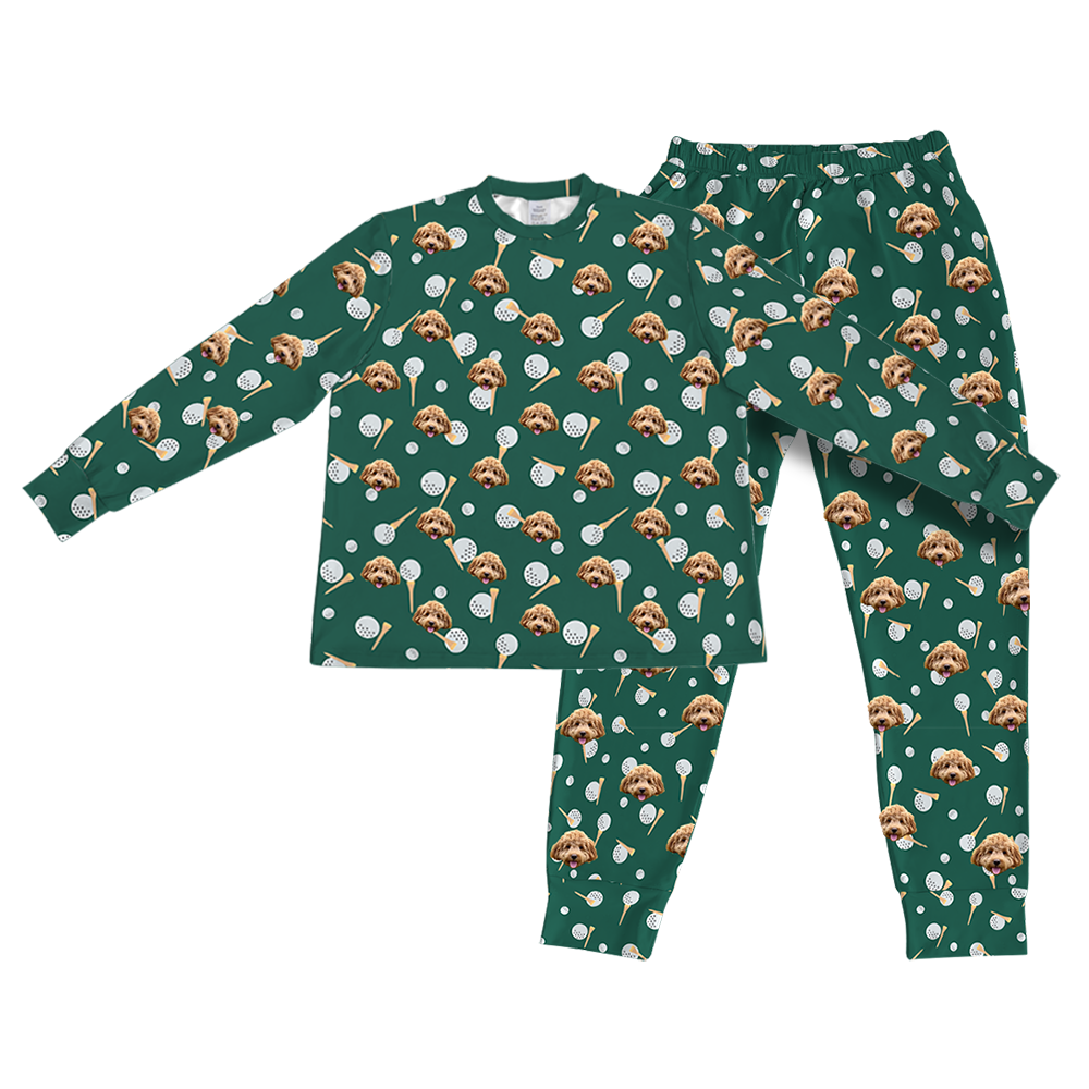 A matching set of long-sleeve pajama top and jogger bottoms, in a dark hunter green fabric. The entire set is covered in a repeating golf-themed pattern featuring alternating elements: white golf balls, light yellow golf tees, and smiling headshots of a brown dog.