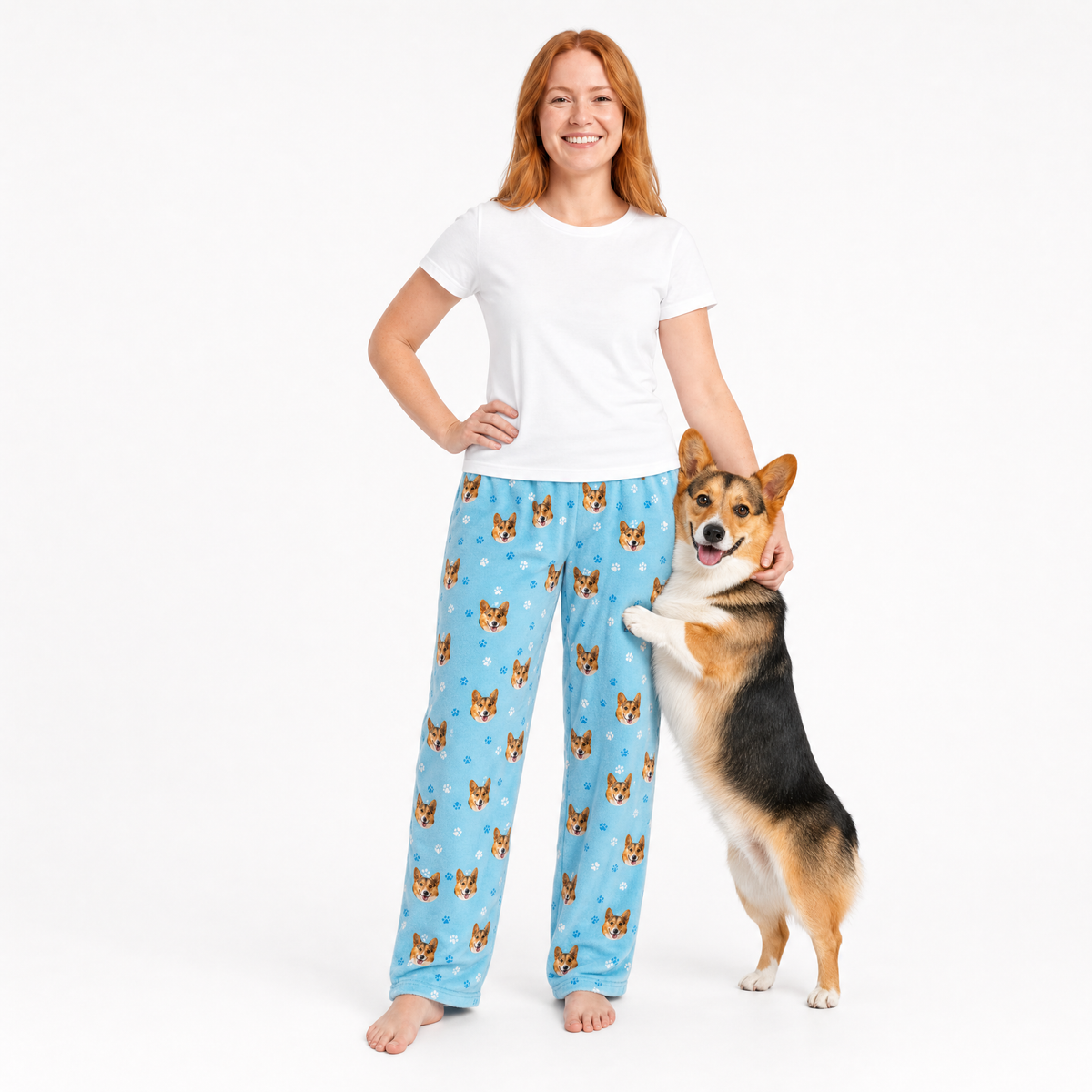 Woman wearing blue pajama pants with dog pattern and a corgi standing next to her on a white background