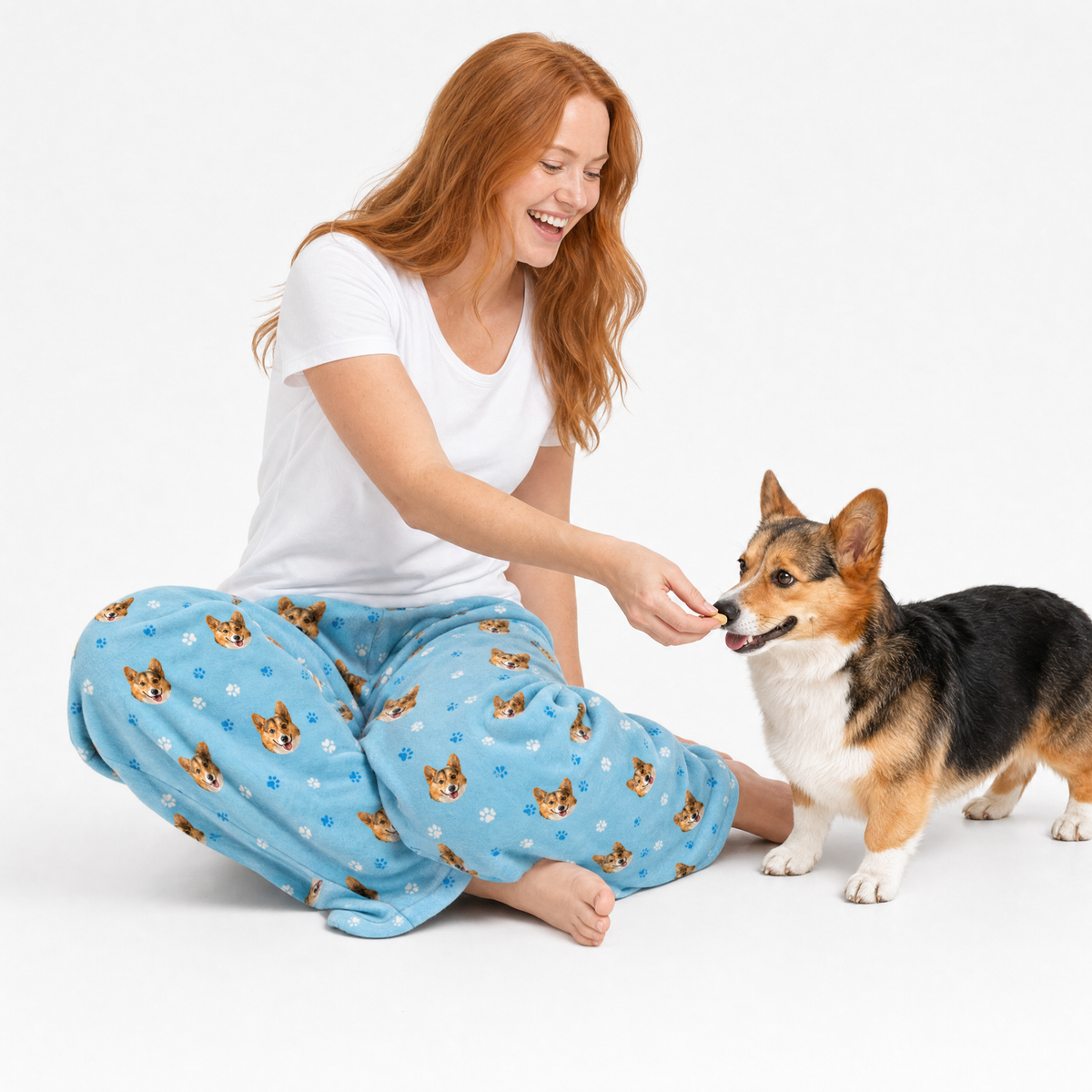 Woman in light blue pajama pants with dog pattern sitting on the floor, interacting with a corgi.