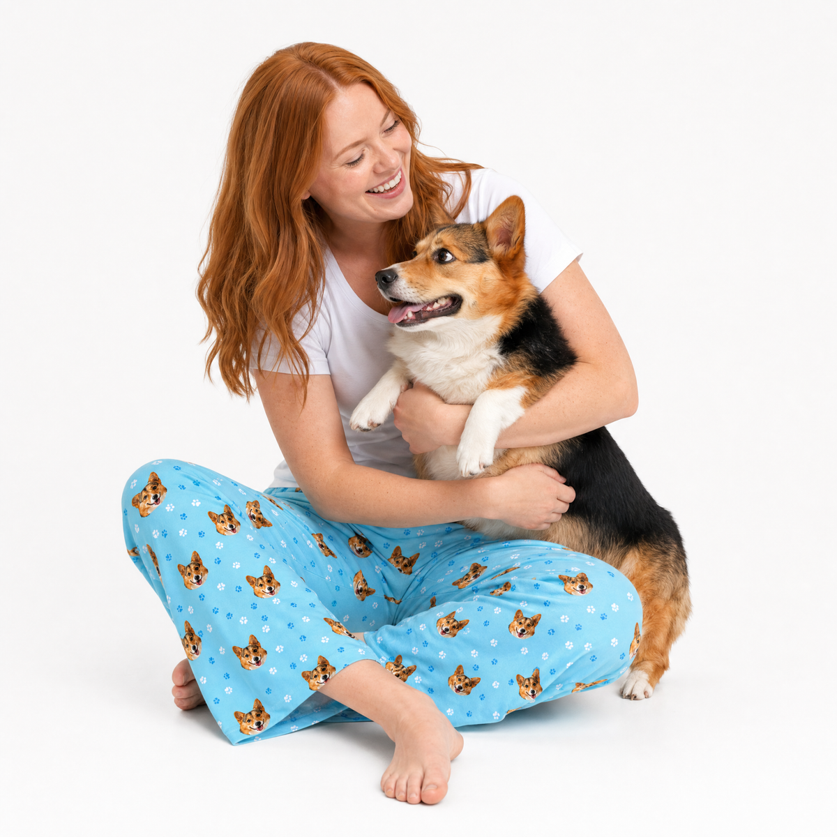 Woman wearing light blue pajama pants with dog pattern holding a corgi on a white background