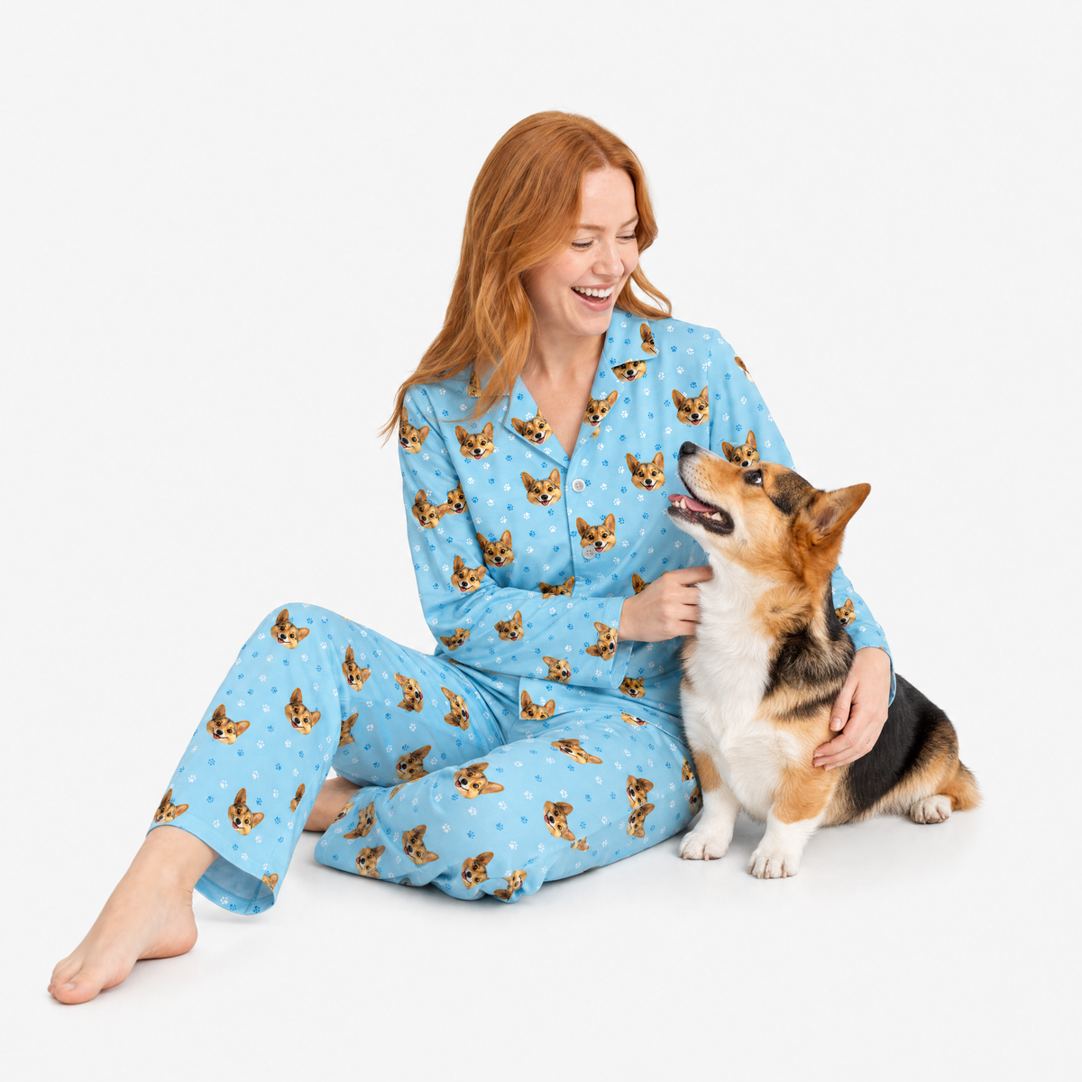 Woman wearing light blue pajamas with dog pattern sitting on white floor with corgi dog.