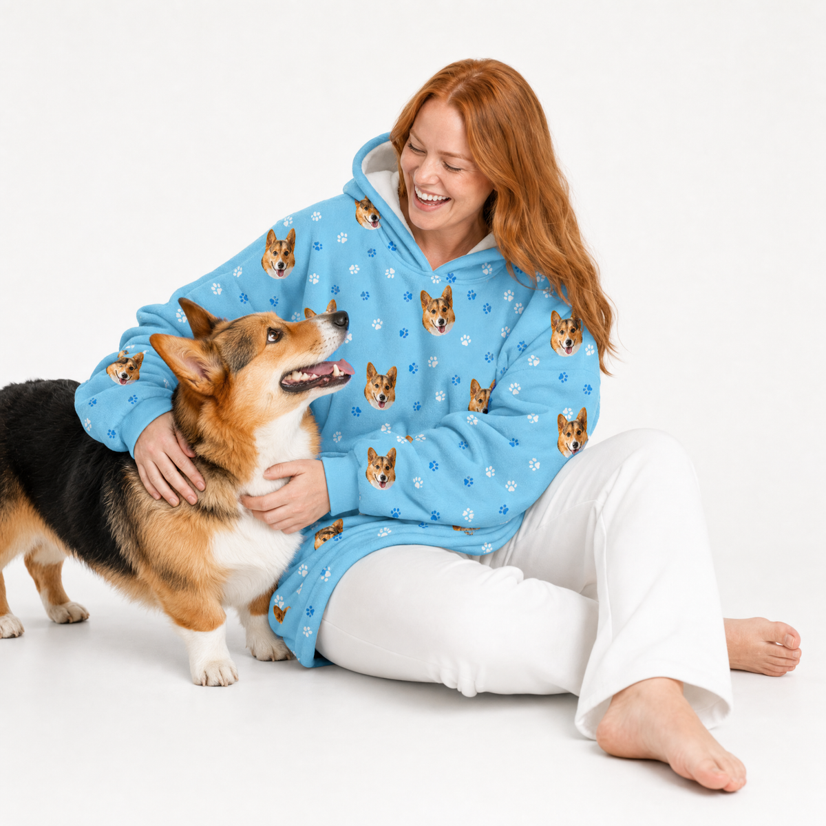 Woman wearing a light blue blanket hoodie with dog patterns, sitting with a corgi on a white background