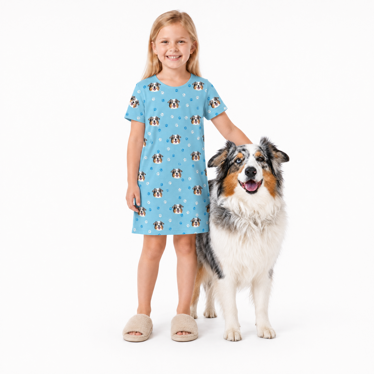 Girl in a light blue nightgown with dog pattern standing next to a dog on a white background