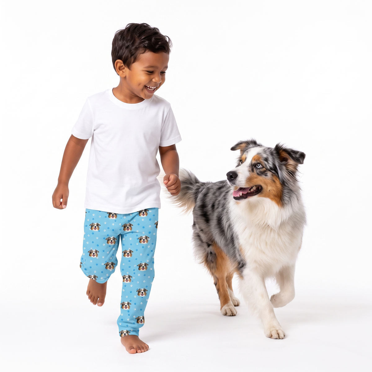 A young boy walking barefoot next to an Australian Shepherd dog, wearing a plain white t-shirt and light blue pajama pants with a dog print, on a solid white background.