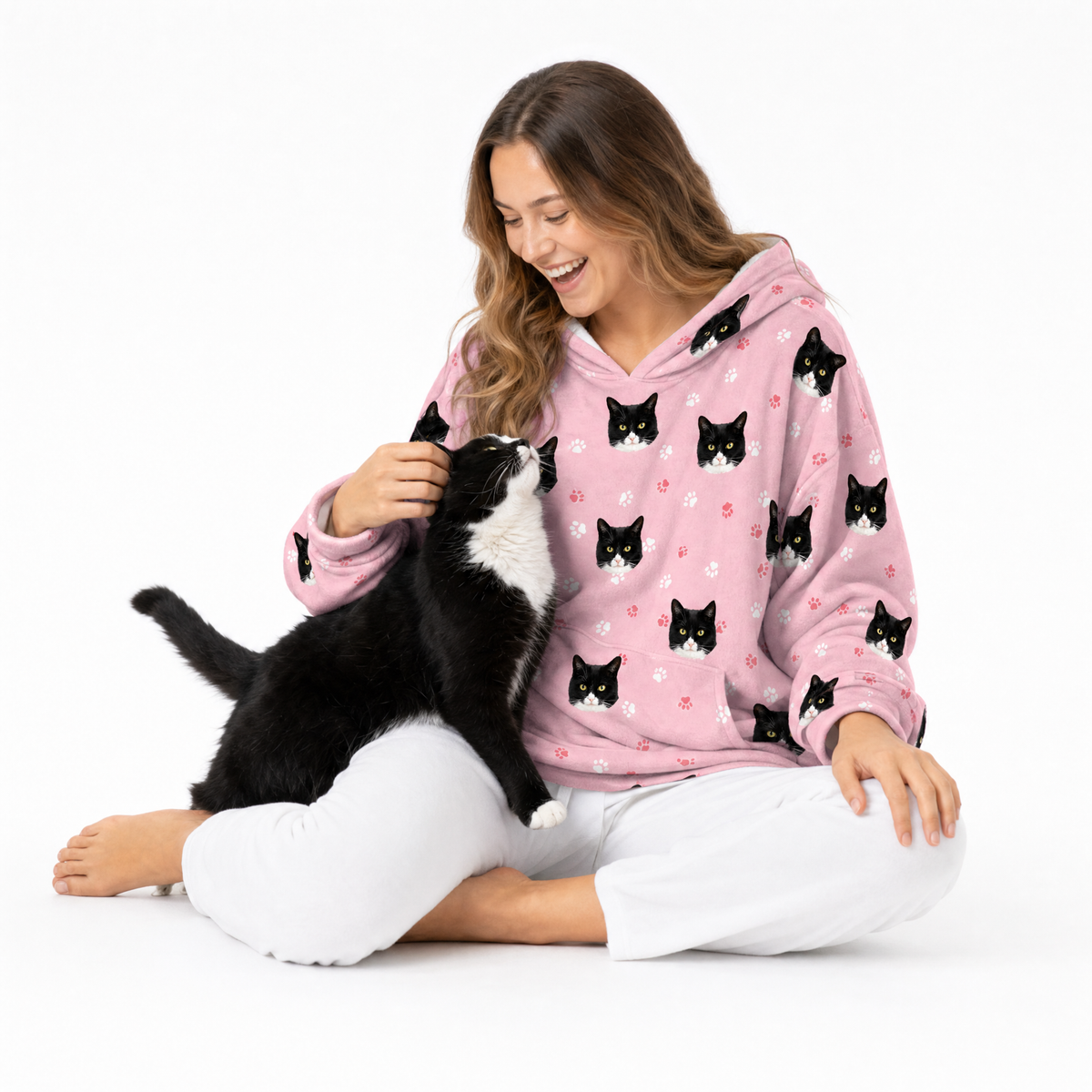 Woman wearing a pink blanket hoodie with black cat patterns, sitting on the floor with a black and white cat.