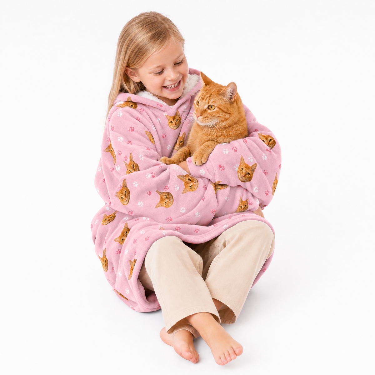 Child in a pink blanket hoodie with cat pattern holding an orange cat on a white background
