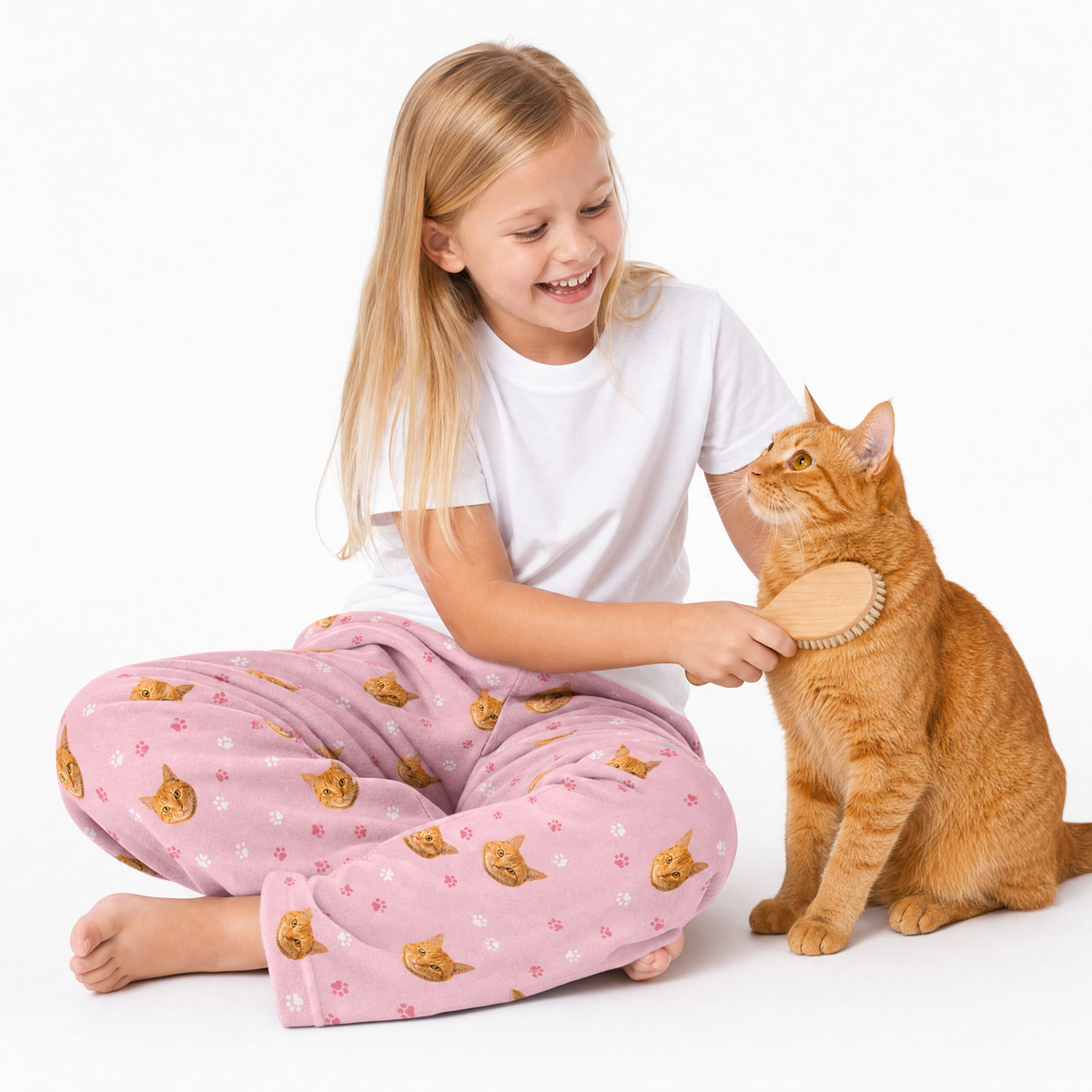 Girl in pink pajama pants with cat pattern brushing a cat on a white background