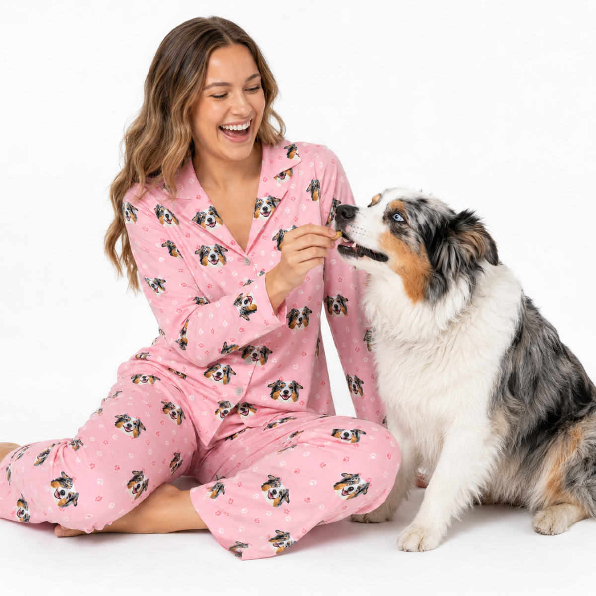 Woman in light pink paws pajamas with dog pattern feeding a dog wearing pink pajamas on a white background