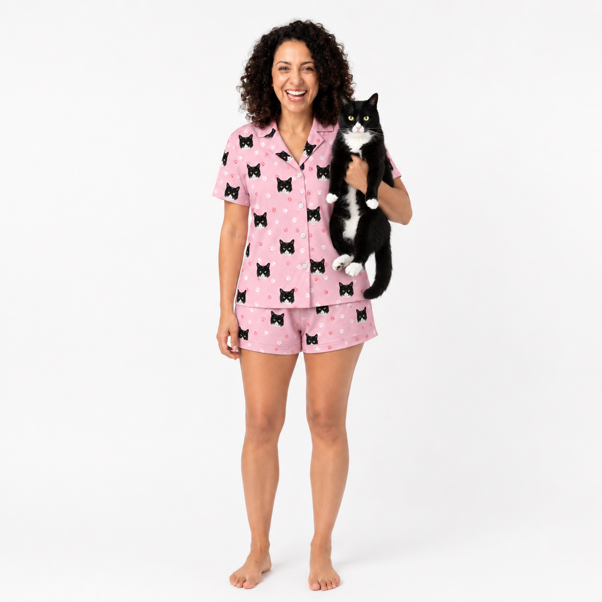 Woman wearing light pink paws pajama set with black cat pattern, holding a black and white cat on a white background