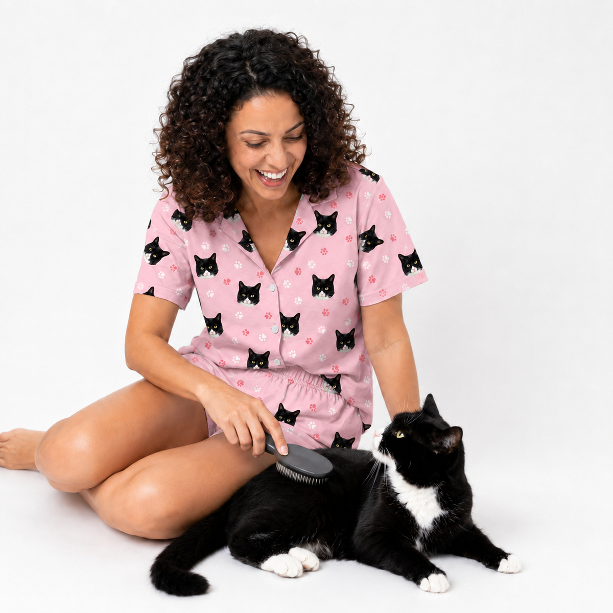 Woman in light pink paws pajamas with cat pattern brushing a black and white cat on a white background
