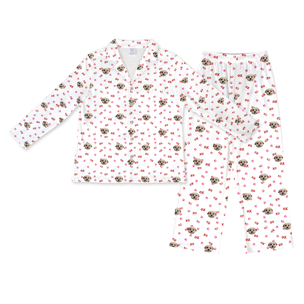 A white long-sleeve button-down pajama set with a repeating pattern. The pattern includes small images of a light brown dog's face, pink "XO" text, and scattered pink paw prints. The set includes a button-down shirt with a collar and matching long pants with an elastic waist.