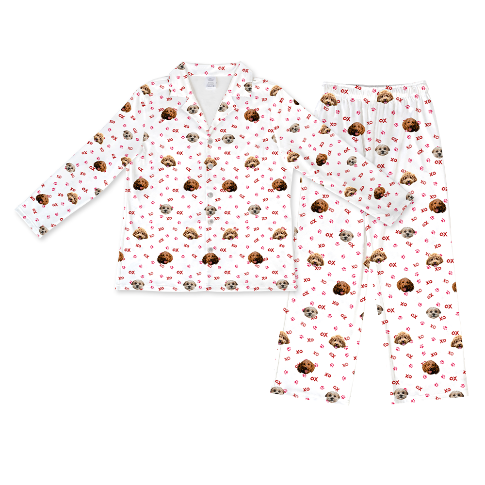 A matching pajama set featuring a long-sleeved, button-down top with a collar and a pair of full-length pants with an elastic waistband. Both pieces are white and display a pattern of two different dogs' faces, along with scattered red "XO" text and red paw prints.