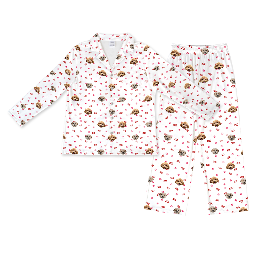 A matching pajama set featuring a long-sleeved, button-down top with a collar and a pair of full-length pants with an elastic waistband. Both pieces are white and display a repeated pattern of a single light brown and white dog's face, along with scattered red "XO" text and red paw prints.