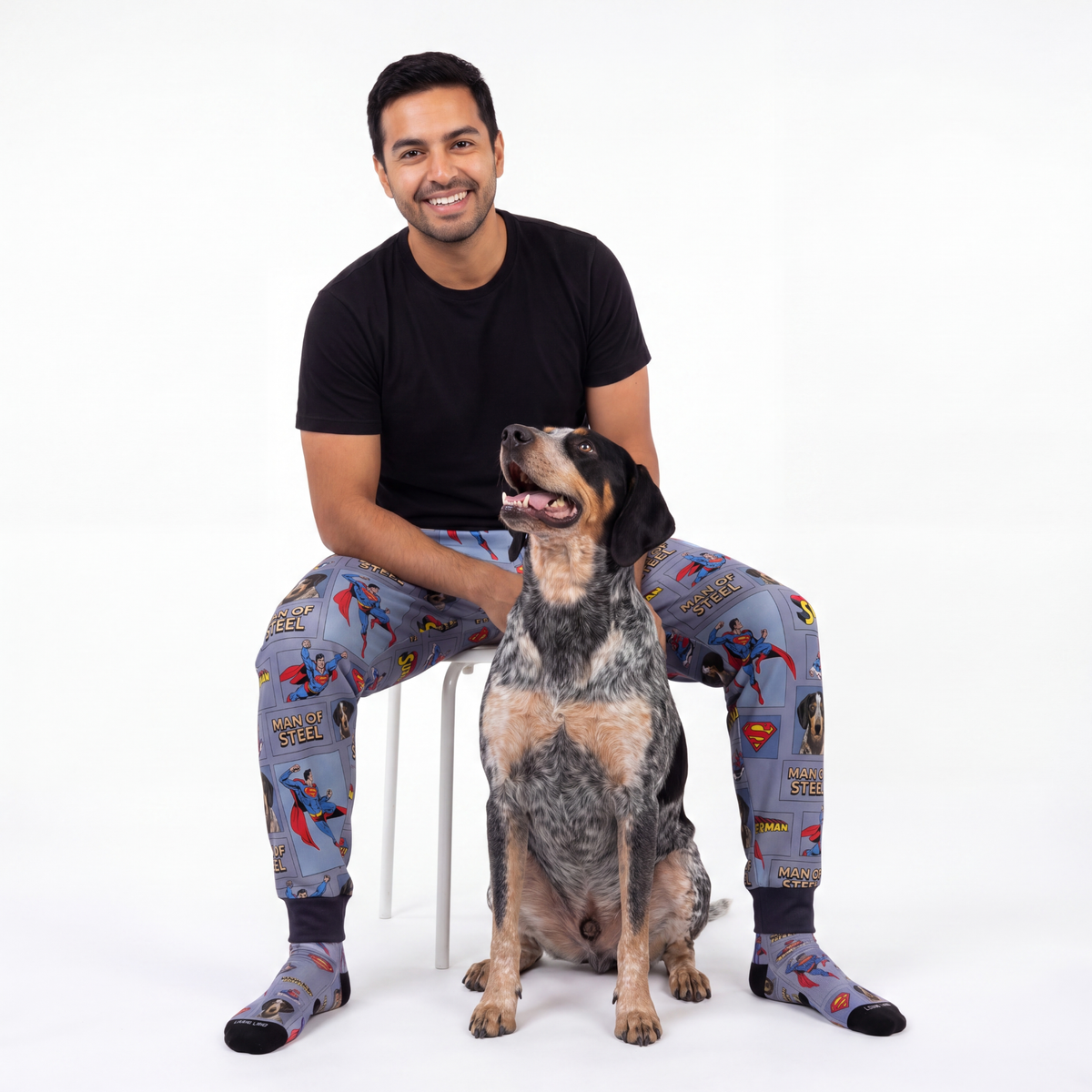 Man wearing superman pajama pants sitting on a chair with a dog.