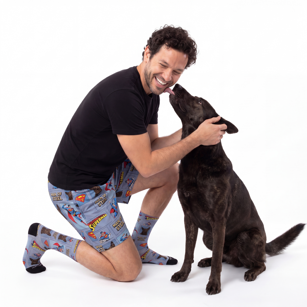 Man wearing superman pajama shorts kneeling with a black dog on a white background