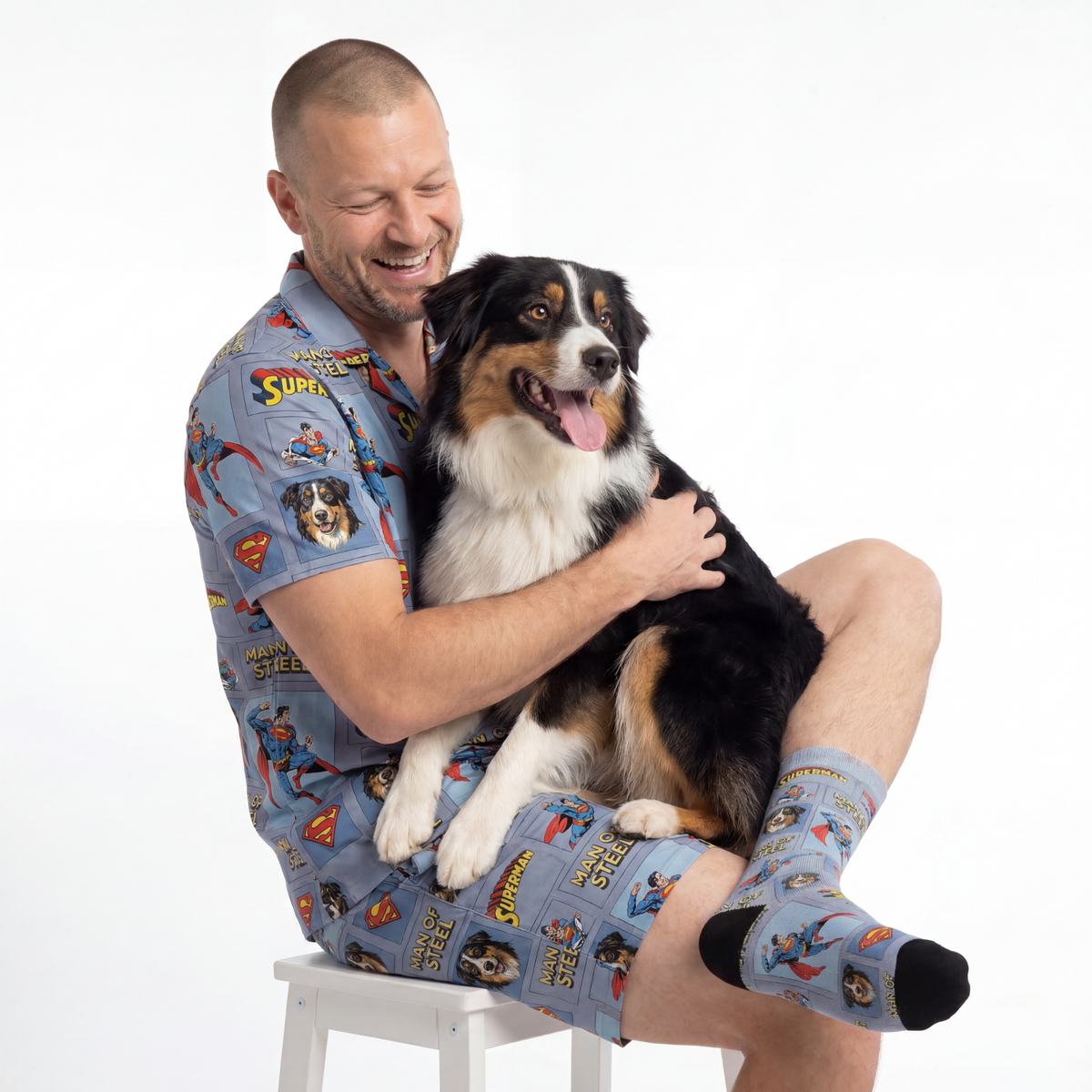 Man sitting on a stool holding a dog, both wearing superman-themed clothing.