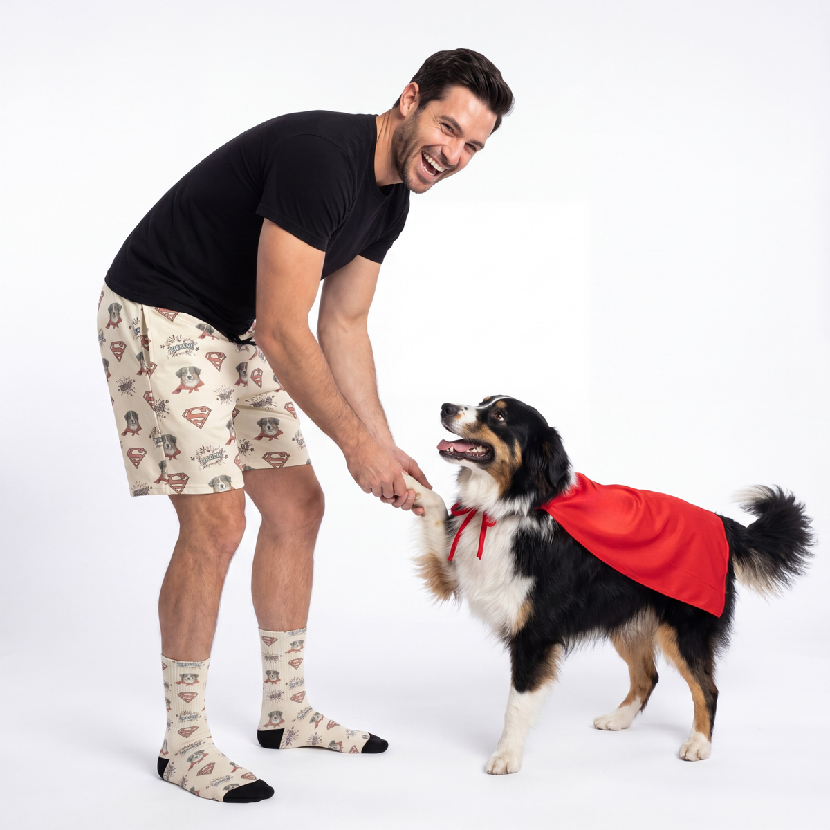Man in wearing Superman shorts interacting with a dog wearing a red cape on a white background
