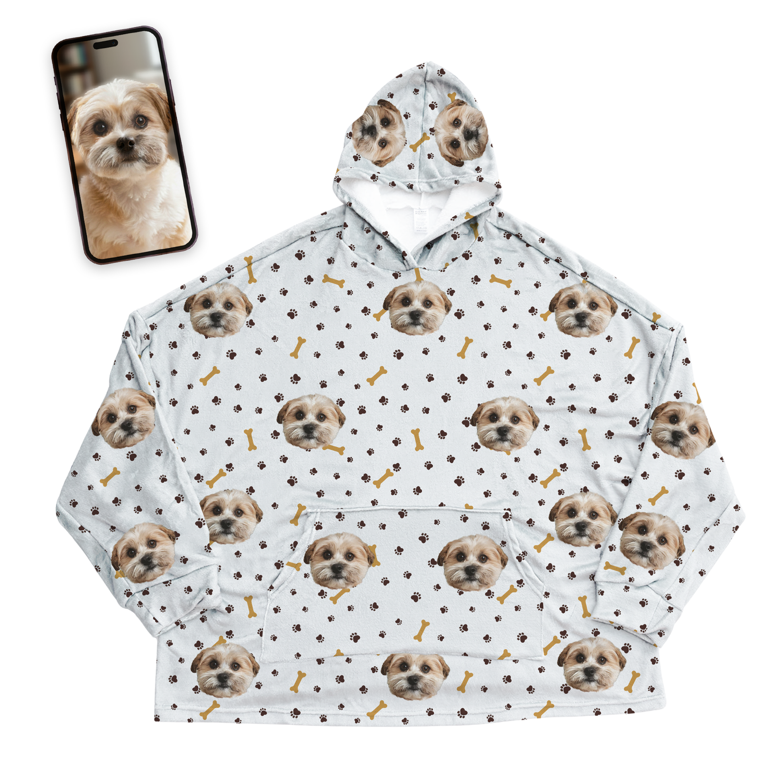 White oversized wearable blanket hoodie decorated with a repeating all-over pattern of a small dog’s face, brown paw prints, and gold dog bones. Shown next to a smartphone displaying the original pet photo used to create the personalized design.