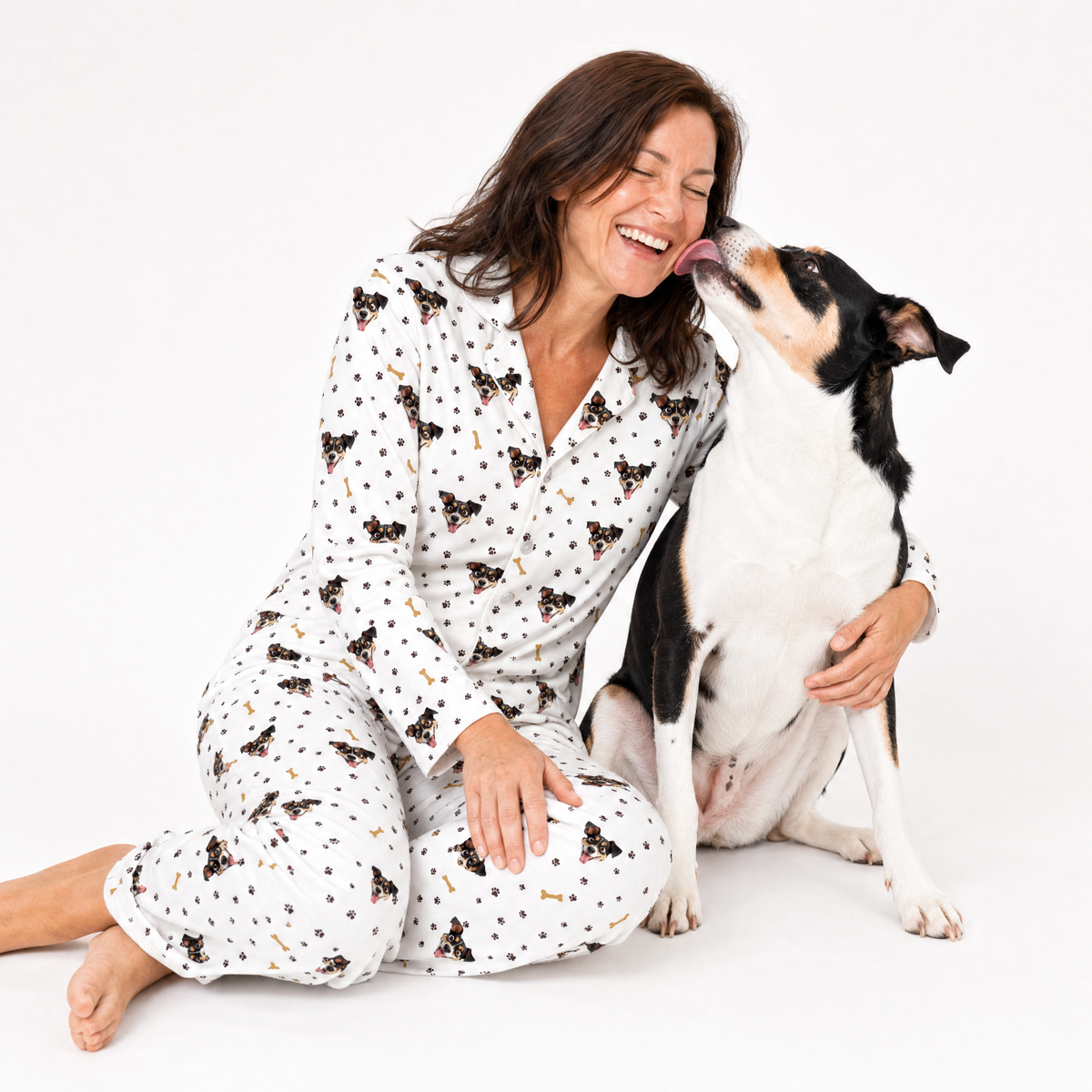 Woman in pajamas sitting on the floor with a dog licking her face against a white background